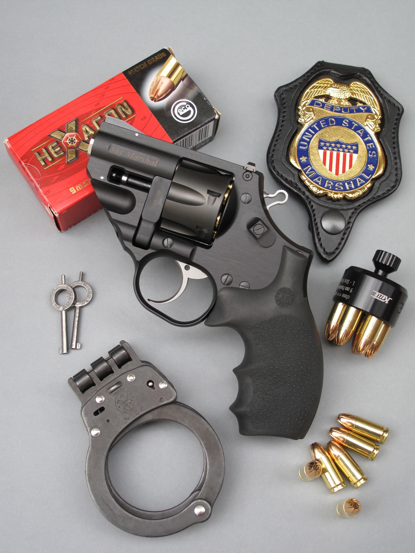 Le revolver Korth Sky Marshal est accompagné ici par son speed-loader, une boîte de cartouches Geco Hexagon de calibre 9 mm Parabellum, une plaque de Deputy U.S. Marchal (police fédérale) et une paire de menottes Smith & Wesson model 300.