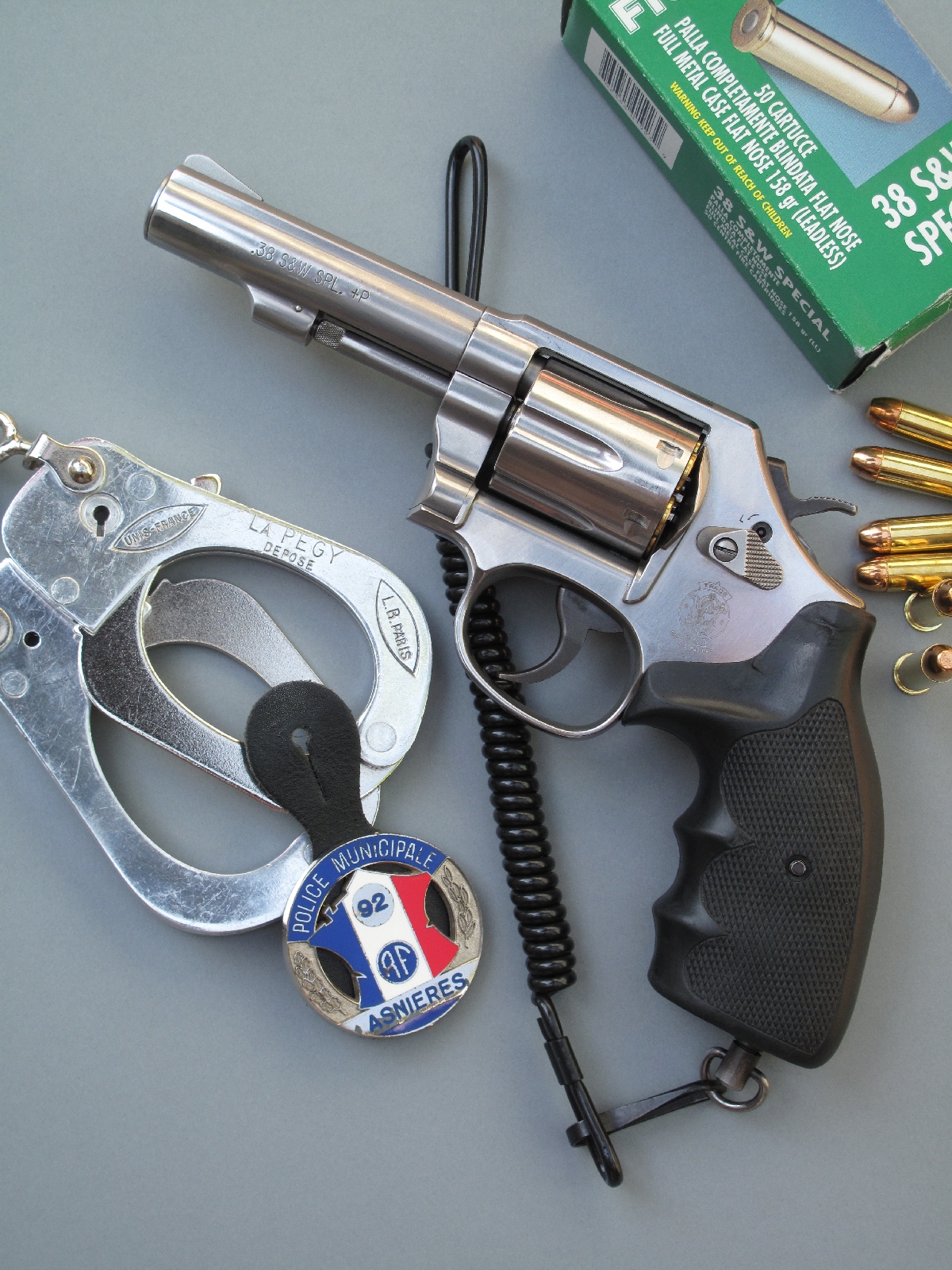 Le revolver Smith & Wesson modèle 64 est accompagné ici d’une paire de menottes, d’un badge de la police municipale et d’une boîte de cartouches de calibre .38 Special manufacturées par la firme italienne Fiocchi.