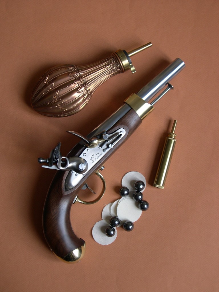 La réplique du pistolet an XIII, réalisée par le fabricant italien Davide Pedersoli, accompagnée ici de balles sphériques de calibre .675 (17,15 mm), en plomb martelé, fabriqués en Allemagne par Haendler & Natermann, de calepins « Black Powder N°1 », en pur coton pour calibre .62 à .69, fabriqués par Pedersoli, d’une poire à poudre en cuivre d’un modèle civil et d’une petite poire d’amorçage en laiton contenant le pulvérin (poudre noire fine PNF4 pour l’amorçage du bassinet).